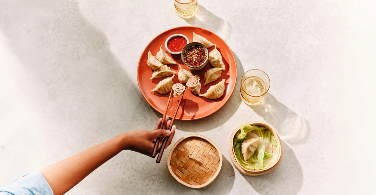 Overhead view of dumplings, dipping sauces and shared dishes on a table with a hand using chopsticks.