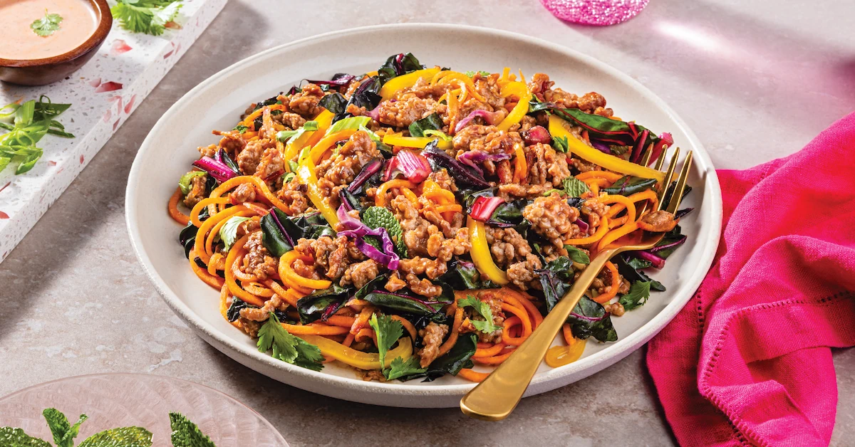 A high-angle close-up of a white plate filled with a colorful stir-fry containing cooked ground pork, orange vegetable noodles, sliced yellow bell peppers, dark leafy greens, purple cabbage, and fresh herbs like cilantro and mint. A gold fork rests on the plate next to a bright pink napkin, with a small wooden bowl of creamy orange sauce in the upper left corner.